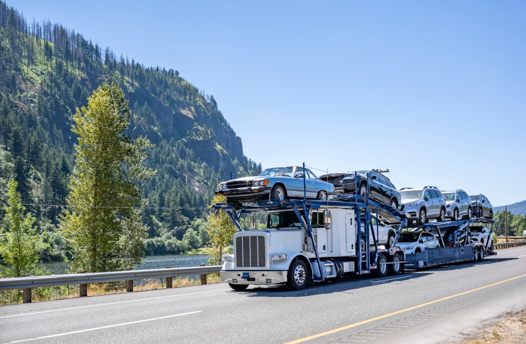 Industrial big rig white classic car shipping hauler semi truck tractor transporting cars on the modular hydraulic two level semi trailer driving on a highway road along the river in Columbia Gorge. 