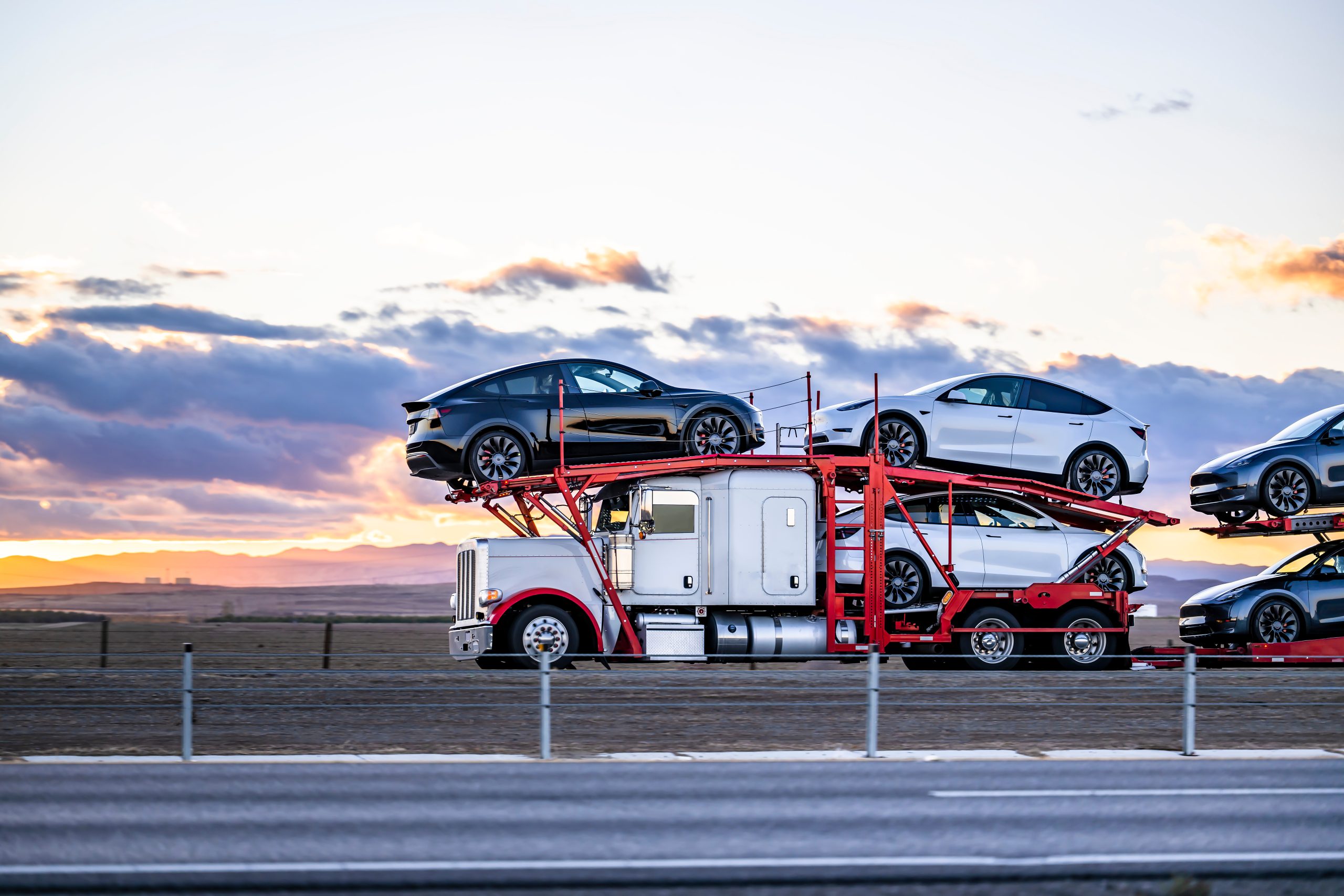 White and red classic car hauler semi truck transporting cars on the hydraulic semi trailer driving on the road at twilight Open transport car shipping from los angeles to houston.
