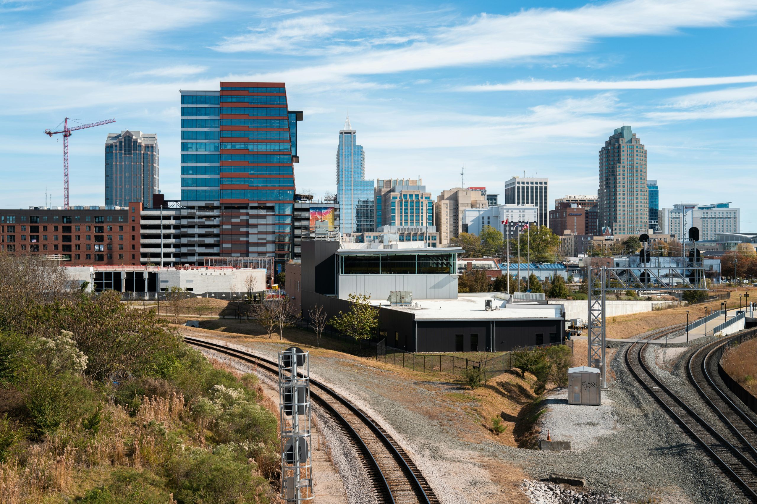 A stunning aerial view of the Raleigh skyline, showcasing modern skyscrapers, surrounding greenery, and a blue sky with wispy clouds.