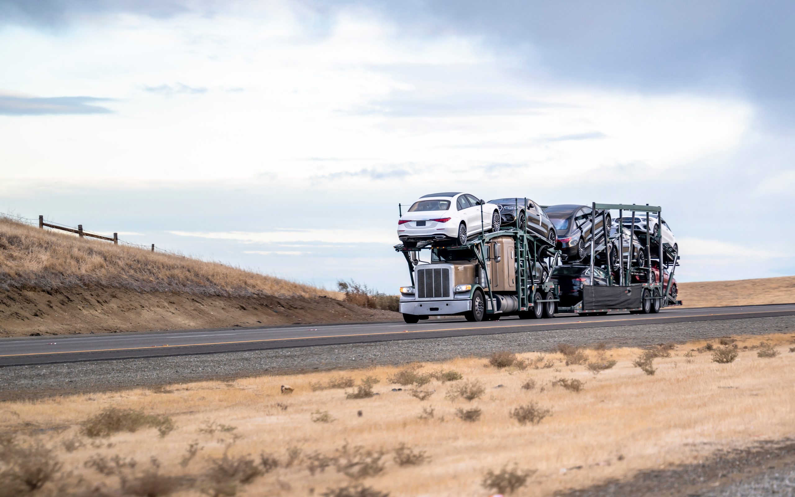 transport-in-carolina Beige commercial big rig car hauler semi truck with turned on headlight transporting fastened cars on two level hydraulic modular semi trailer running on the highway road in south carolina at twilight
